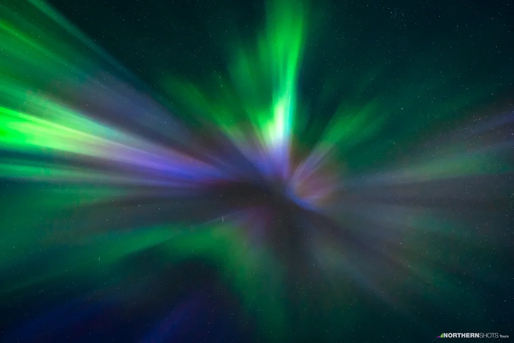 Radiating aurora rays in blue, purple, pink, white, and green fanning out from a central point in the night sky.