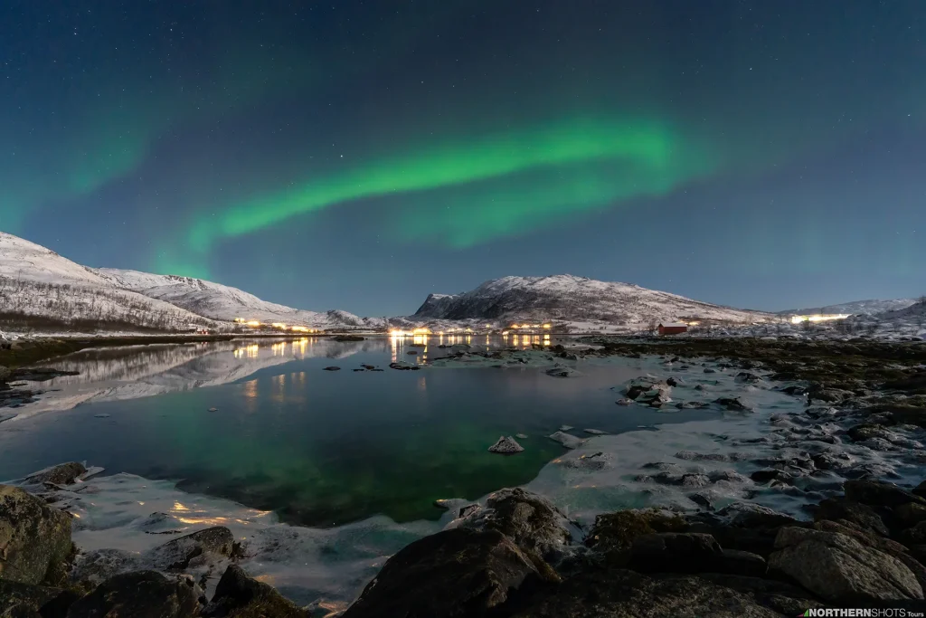 Circular aurora borealis reflected over a moonlit lake with distant house lights along the far shoreline.