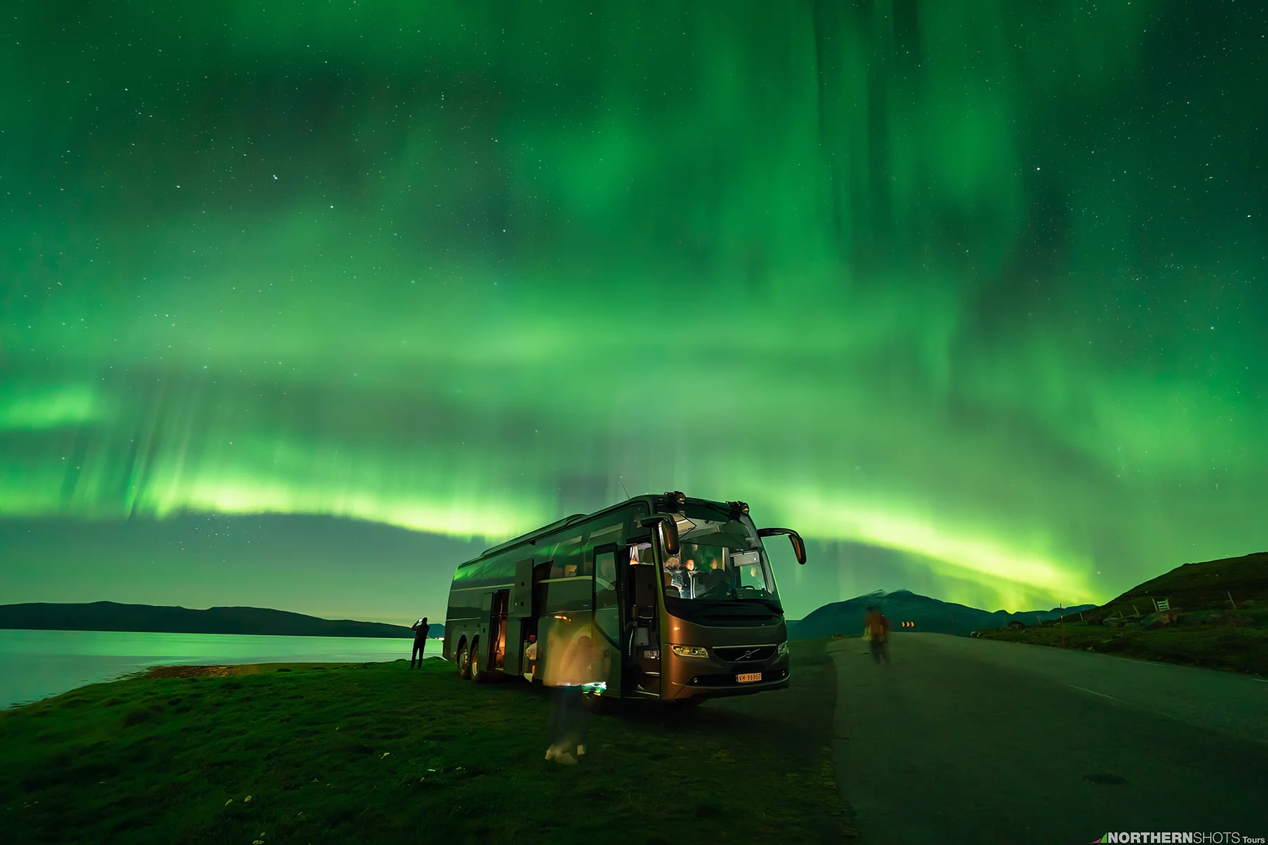 Tour bus parked by a fjord under a wide aurora arc illuminating the shoreline and surrounding landscape.