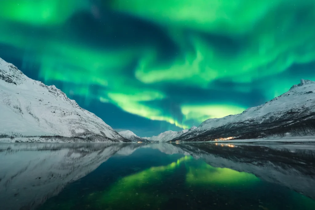 Green aurora clouds mirrored in a still Arctic fjord, surrounded by snowy peaks under the night sky.