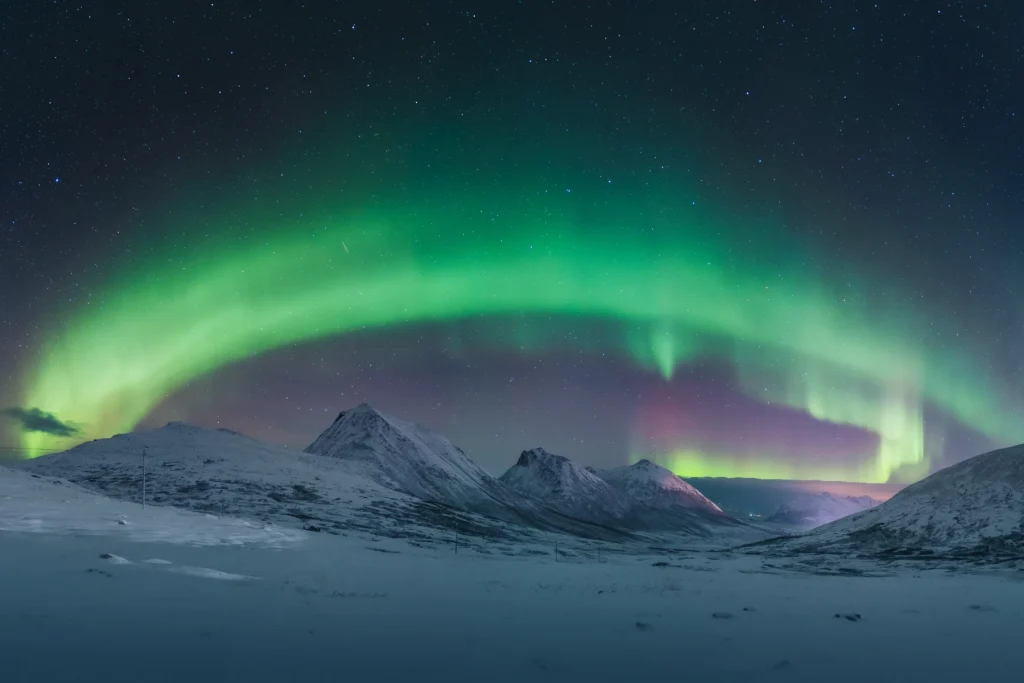 Aurora borealis shaped like a rainbow in the night sky, arching in purple and green above snow-covered mountain peaks.