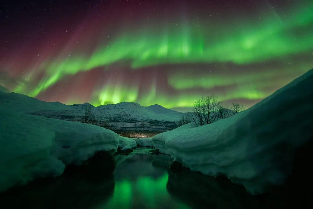 Low-angle view of colourful Northern Lights over a snow-covered lakeshore, with distant mountain peaks under the night sky.