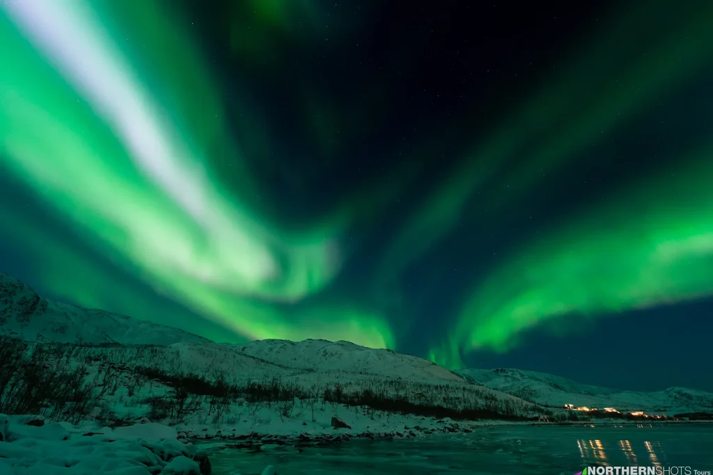 Blazing green and white Northern Lights arc over snowy mountain peaks as seen from the edge of a frozen fjord.