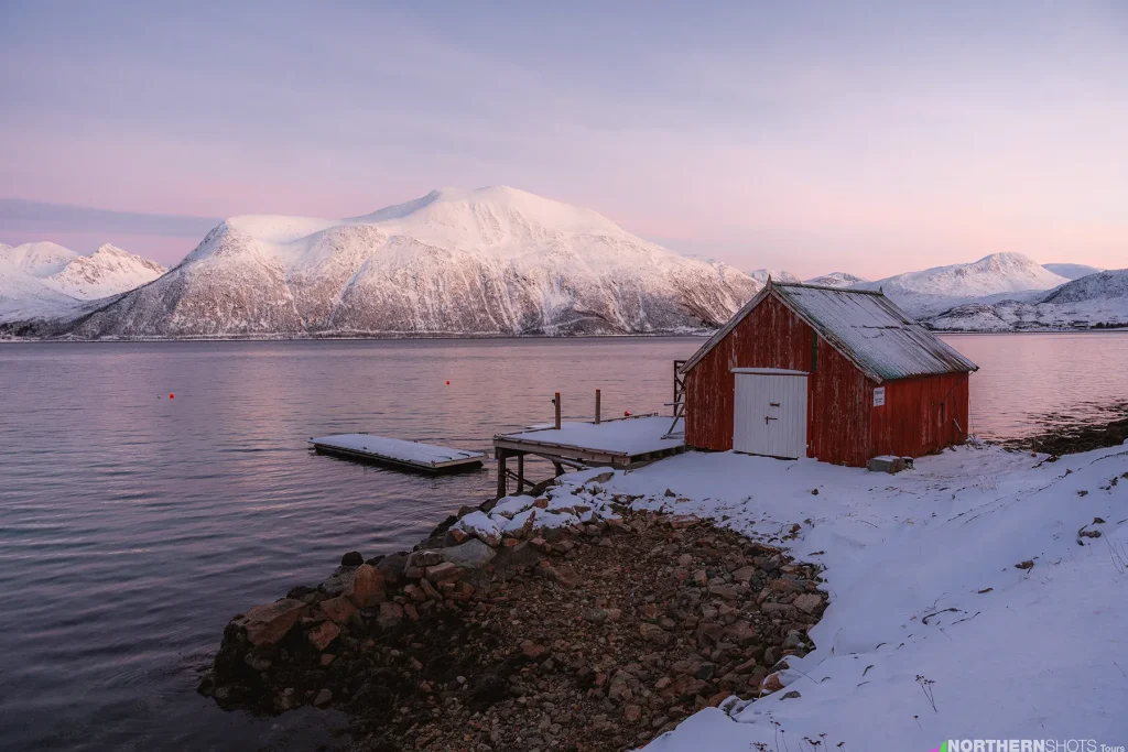 Scenic view of a coastal boathouse and pier overlooking a fjord surrounded by snow-covered mountains on Kvaløya near Sommarøy.
