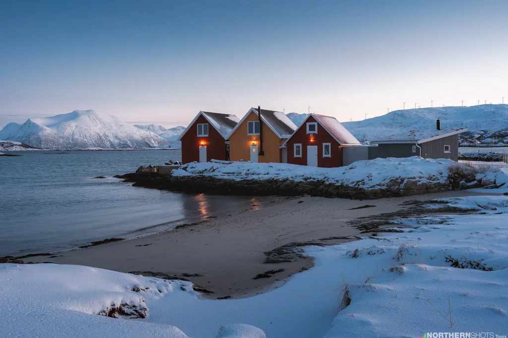 Colourful cabins along Sommarøy’s shoreline under soft Arctic light, with snowy peaks in the background.