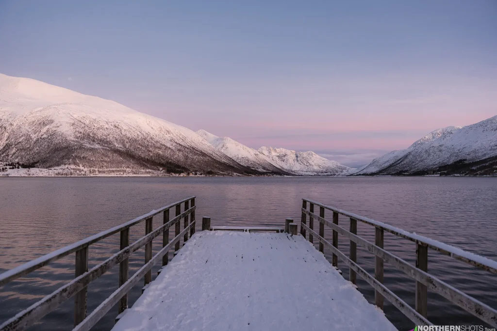 View from a snowy pier reaching into a fjord around Kvaløya, where distant mountains are lit by gentle pink twilight.