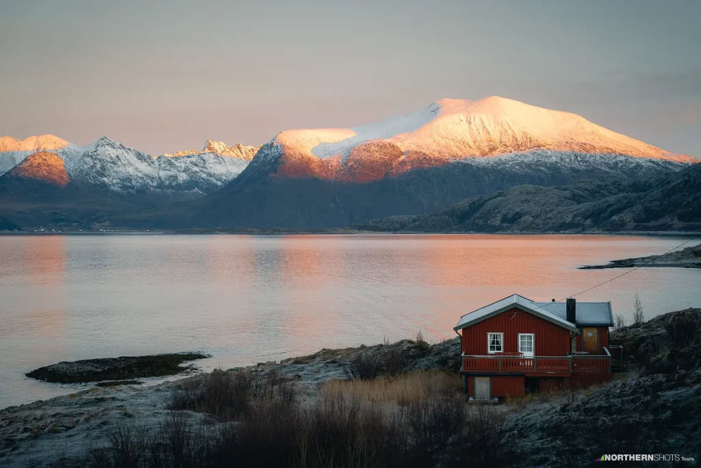 Red cabin on Kvaløya’s rocky shoreline near Sommarøy, with distant snowy mountains lit by low Arctic sunlight.