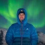 NorthernShotsTours guide Marek in winter gear and headlamp, standing against a blurred backdrop of auroras over snowy Arctic peaks.