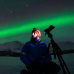 Tour guide Michele kneeling in the snow with his tripod as the Northern Lights glow behind him during an Aurora Chase.