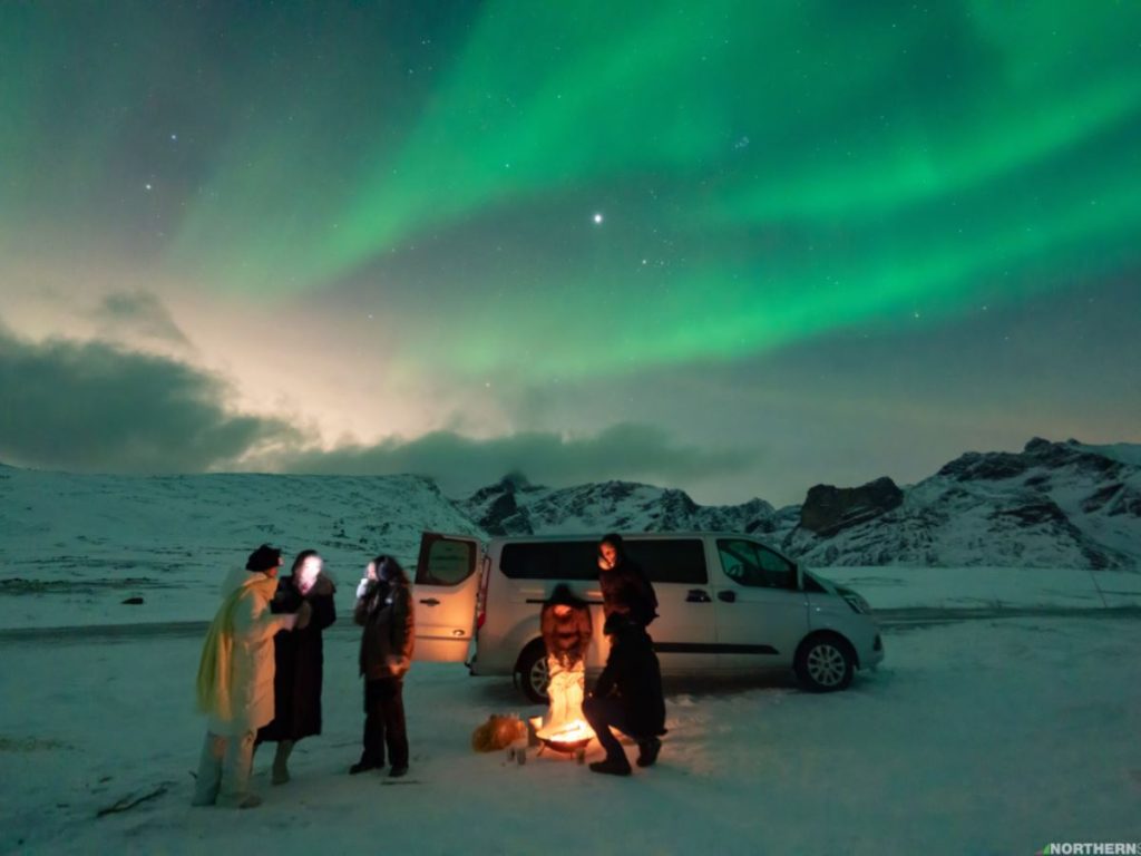 Guests gathered by a campfire in front of their van with a massive aurora display and moonlight above.