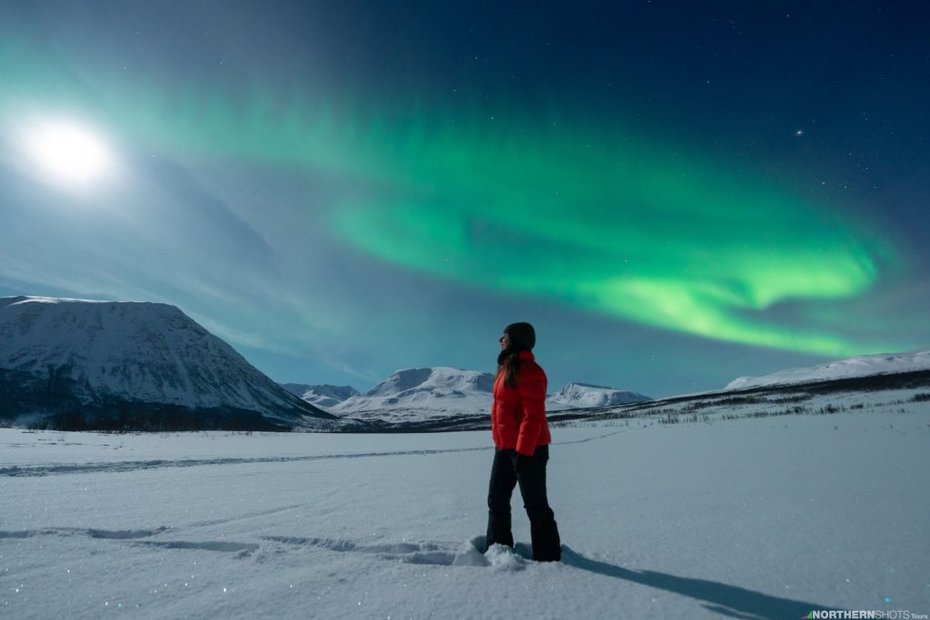 Woman standing sideways in the snow, gazing toward the full moon over the mountains, with a sweeping aurora behind her stretching into the horizon.