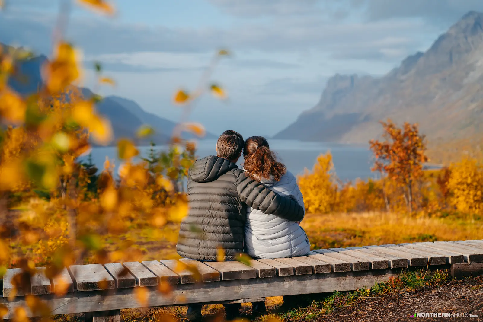 Couple sitting on wood structure, embracing while overlooking a fjord surrounded by autumn-coloured trees.