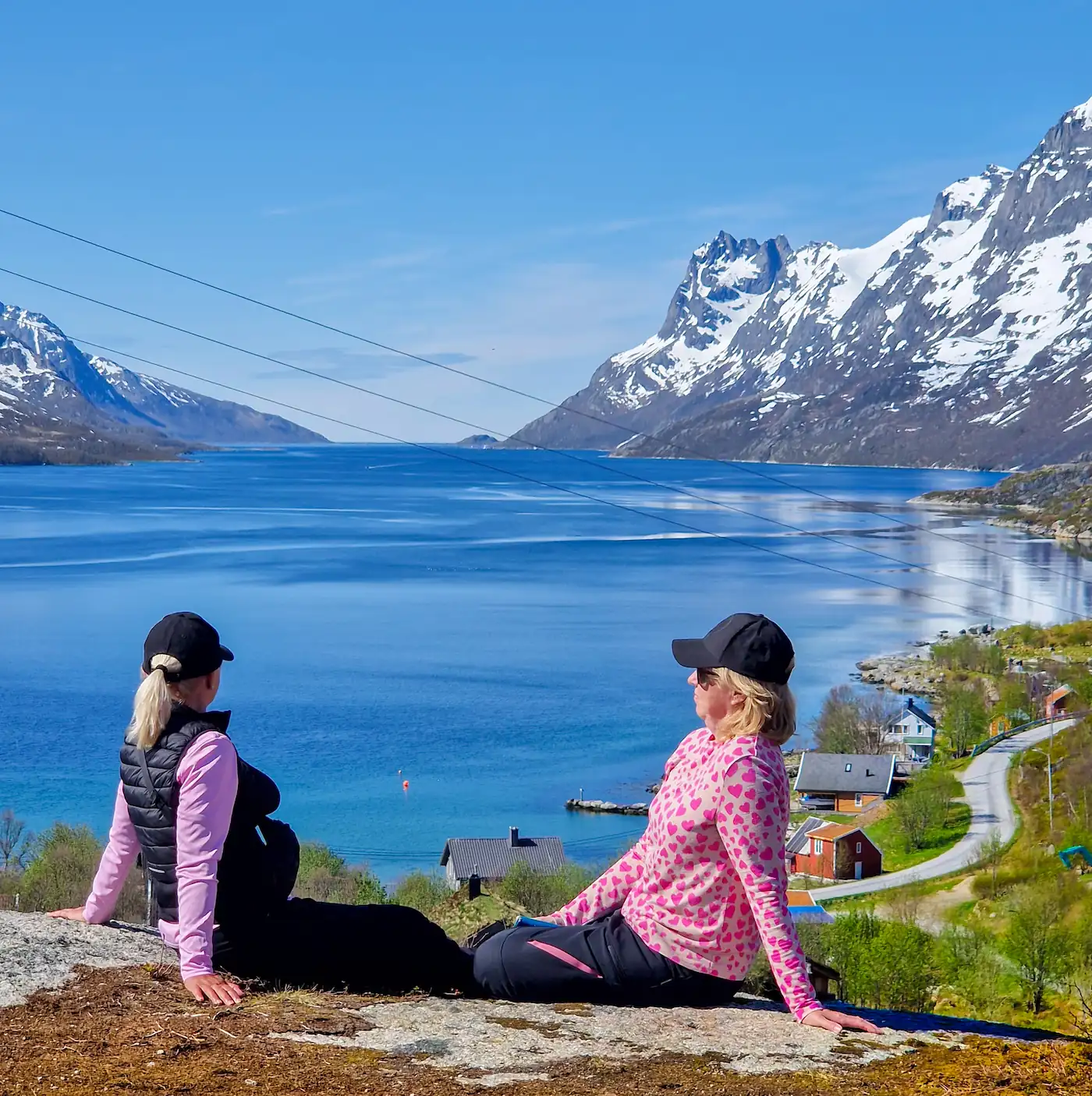 Tourists relaxing on mountain top near Tromsø, overlooking fjord with snowy peaks in summer.